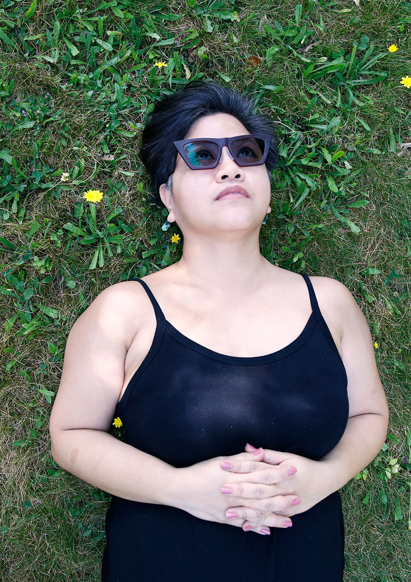 Photograph of essay author Fran Flaherty, a Filipino woman wearing sunglasses and a black jumpsuit. She's lying in the grass with small yellow wildflowers around her.