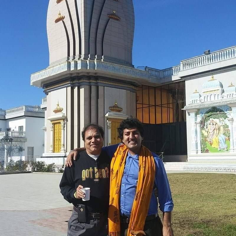 Photograph of two Brown men stand with their arms around each other in front of a Hindu temple. Both are facing the camera. Puneet stands on the right with a big smile on his face.