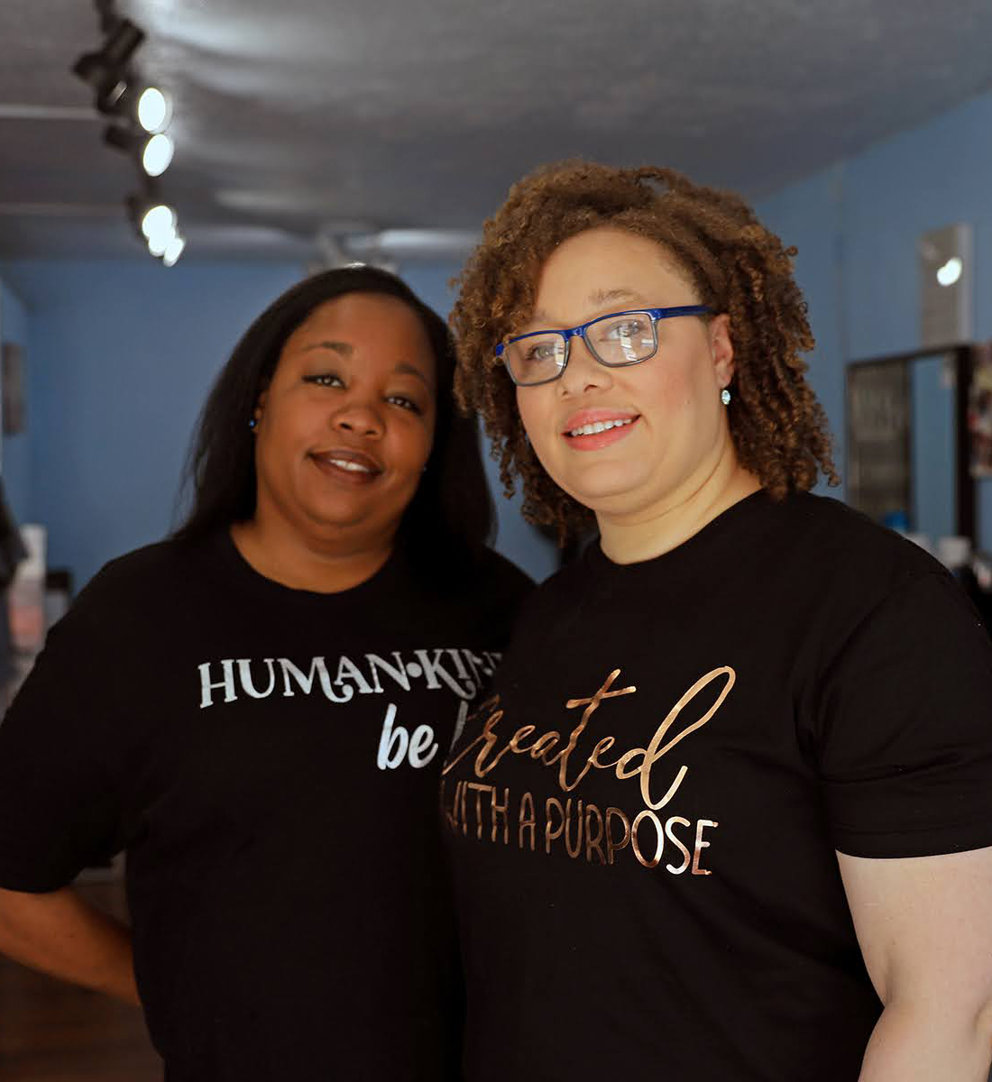 Two Black women smile at the camera and stand side by side in a beauty studio. One wears a shirt that says 'humankind be kind' and the other's shirt says 'created with a purpose'.